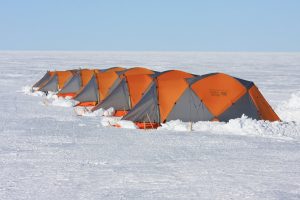A row of orange and gray tents is set up on a flat, snowy landscape under a clear blue sky, suggesting an Antarctic Logistics and Expeditions campsite in the remote wilderness of Antarctica.