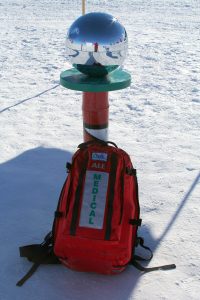 A red medical backpack labeled MEDICAL rests on snow beneath a shiny metallic sphere atop a pole at the South Pole, reflecting the Antarctic surroundings and a person taking the photo—an iconic scene of Antarctica Logistics and Expeditions.