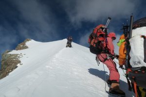 Several climbers in colorful gear ascend a steep, snow-covered mountain ridge in Antarctica under a partly cloudy sky, using ropes and helmets, with rocky peaks visible in the distance—a true Antarctic Logistics and Expeditions adventure.