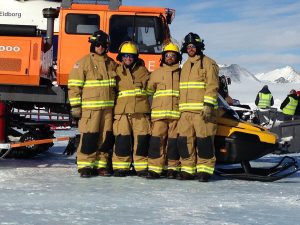 Four people in tan firefighting gear and helmets stand together on ice at Antarctica, in front of an orange vehicle and snowmobile, with snowy mountains, other people, and Antarctic Logistics and Expeditions support in the background.