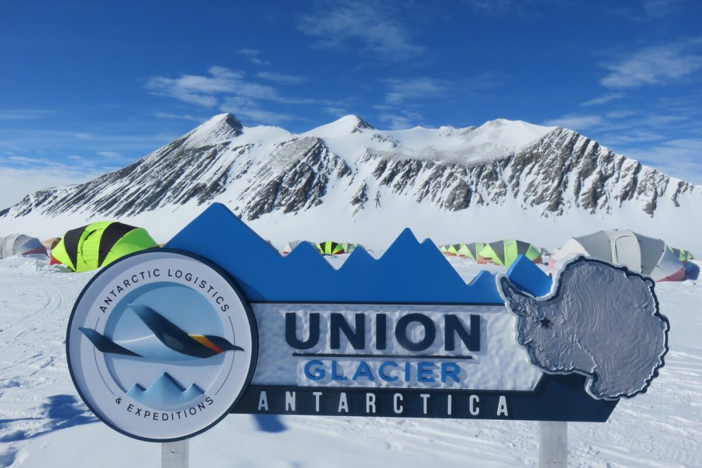 A sign reading “Union Glacier, Antarctica” stands in snowy terrain with snow-capped mountains and colorful tents in the background under a bright blue sky, marking a hub for Antarctic Logistics and Expeditions near the South Pole.