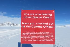 A red sign in snowy Antarctica reads: You are now leaving Union Glacier Camp. Have you checked out at the Comms Office? Additional safety instructions and the Antarctic Logistics and Expeditions logo stand against a backdrop of snowy mountains.