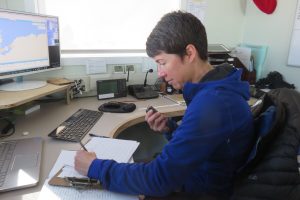 A person in a blue jacket sits at a desk at Antarctic Logistics and Expeditions, holding a radio and writing on papers. A monitor displays a map of Antarctica, with various office equipment including a laptop, phone, and clipboard nearby.