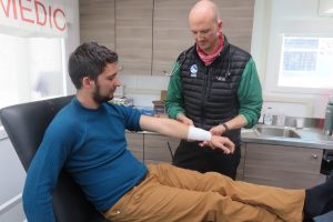 A medic with Antarctic Logistics and Expeditions wraps a bandage around a seated man's forearm in a South Pole clinic. The man rests his arm on his lap while the medic checks the bandage, with medical supplies visible in the background.