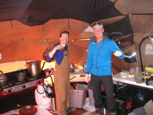 Two people smile inside a tent kitchen at the South Pole, holding drinks. Surrounded by cooking equipment and supplies, they wear warm outdoor clothing as snow covers the floor—an authentic Antarctic Logistics and Expeditions experience.