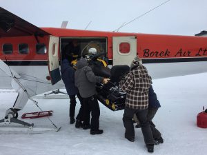 Four people on snow in Antarctica load a snowmobile into a small red and white Antarctic Logistics and Expeditions aircraft, with another person inside helping guide the vehicle.