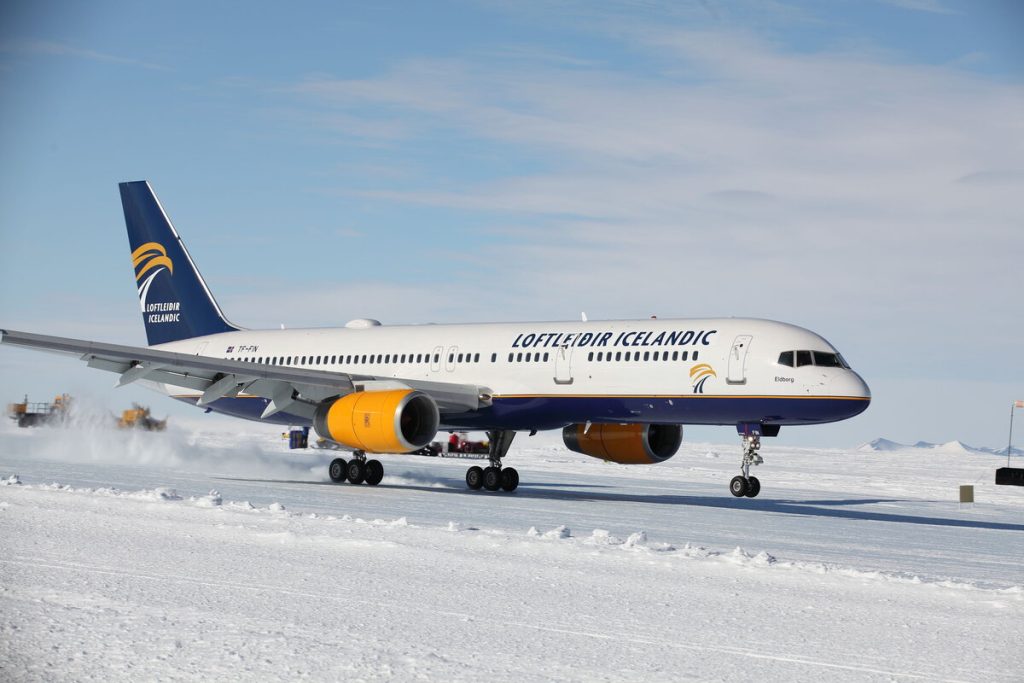 An Icelandair plane, painted white, blue, and yellow, taxis on a snowy runway under a clear sky in Antarctica. The airline logo and “Loftleidir Icelandic” text are visible on the fuselage, supporting Antarctic Logistics and Expeditions near the South Pole.