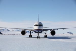 A commercial airplane operated by Antarctic Logistics and Expeditions is parked on a snow-covered runway near the South Pole, with majestic mountains in the background under a clear blue sky.