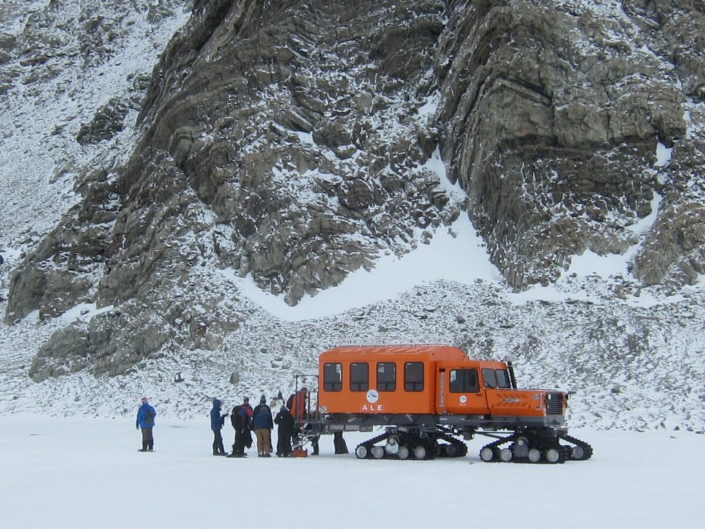 A group of people stand beside an orange tracked vehicle labeled ALE (Antarctic Logistics and Expeditions) on a snowy, frozen landscape in Antarctica, with rocky, snow-dusted cliffs in the background.
