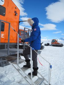 A person in a blue parka with a fur-lined hood climbs metal stairs into an orange vehicle on a snowy Antarctic landscape, with snowcats and mountains visible in the background under a partly cloudy sky at the South Pole.