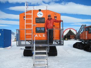 A person in a red jacket stands on the steps of a bright orange Antarctic Logistics and Expeditions tracked vehicle, surrounded by snow and equipment in an Antarctica polar landscape.