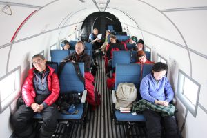 Passengers sit and rest in blue seats inside a small, narrow airplane with white walls. Some people, headed for Antarctica with Antarctic Logistics and Expeditions, look out the windows, read, or sleep as bags and jackets are scattered on empty seats and the aisle floor.