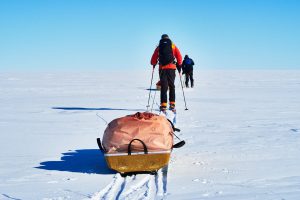 Two people in winter gear ski across a vast, snowy Antarctic landscape, pulling a sled loaded with supplies under a clear blue sky—capturing the spirit of South Pole expeditions.