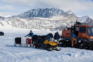 People in cold-weather gear stand beside a yellow snowmobile, sled with tanks, and an orange tracked vehicle on a snowy landscape in Antarctica, with rugged mountains beyond—part of an Antarctic Logistics and Expeditions journey near the South Pole.