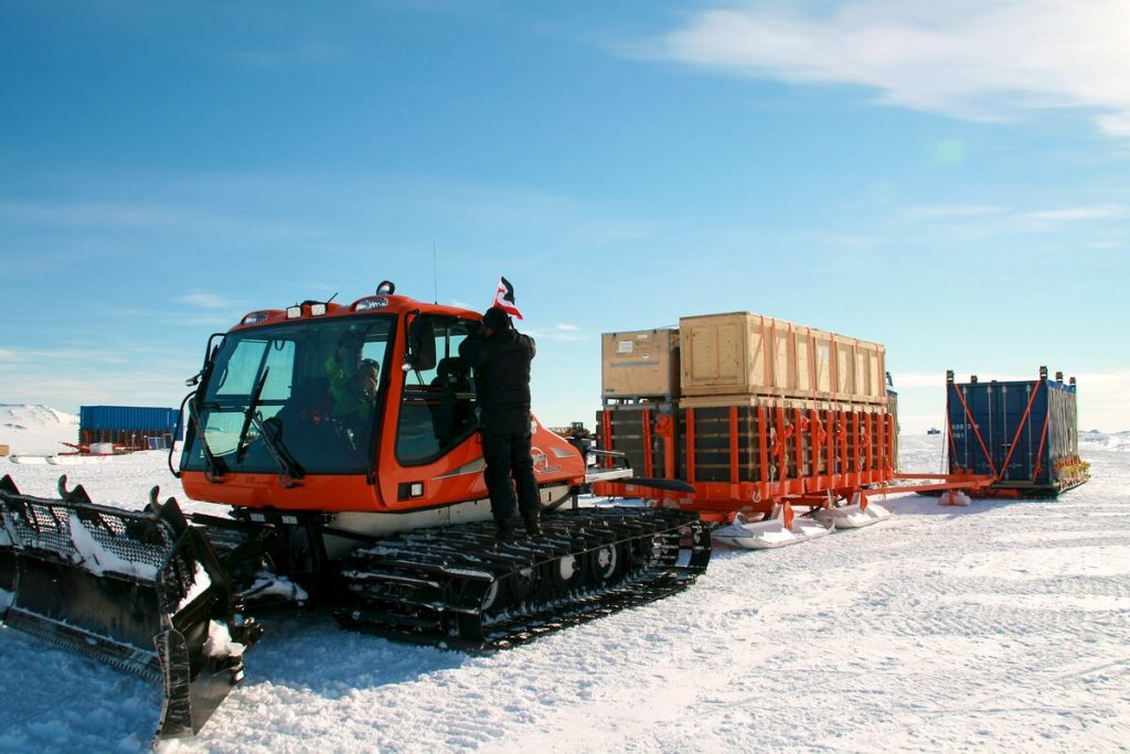 An orange snowcat vehicle pulls sleds loaded with large crates and containers across the snowy landscape of Antarctica under a clear blue sky; a person stands beside the vehicle, supporting Antarctic Logistics and Expeditions near the South Pole.