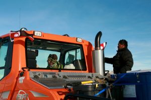 Two people in winter clothing stand by an orange Antarctic Logistics and Expeditions snow vehicle under a blue sky near the South Pole; one sits inside the cab, the other stands outside near the exhaust pipe. Small red-and-white flags are attached to the vehicle.