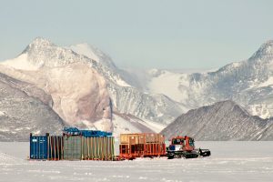 A snow tractor pulls several large sleds loaded with cargo containers across a snowy, flat Antarctic landscape, with rugged, snow-covered mountains in the background.