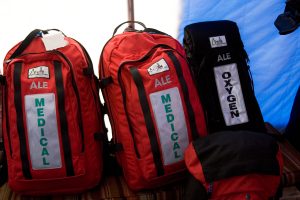 Three red and black medical backpacks labeled ALE MEDICAL and one black bag labeled ALE OXYGEN stand against a blue tent wall at an Antarctic Logistics and Expeditions site. A smaller backpack lies in front of them.