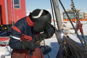 A person with winter gear and a welding helmet welds metal outdoors in a snowy South Pole environment, sparks flying as industrial equipment stands in the background—capturing the challenges faced by Antarctic Logistics and Expeditions teams.