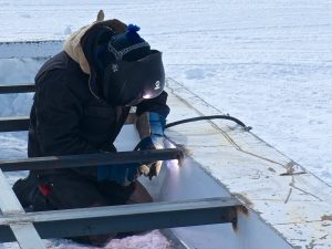 A person wearing a welding mask and gloves welds metal beams outdoors on snow at the South Pole, with visible sparks and protective gear—showcasing the challenging conditions of Antarctica.