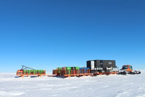 A tracked vehicle pulls orange sleds carrying fuel drums and a black research container across the flat, snowy expanse of Antarctica under a clear blue sky, supporting South Pole operations for Antarctic Logistics and Expeditions.