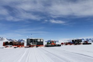 Several sleds carrying equipment, containers, and a small building are being pulled across a snowy, mountainous landscape in Antarctica under a partly cloudy sky, likely as part of an Antarctic Logistics and Expeditions research mission.