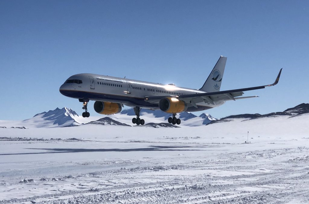 A large commercial airplane with yellow engines, operated by Antarctic Logistics and Expeditions, is landing on a snowy, icy runway near the South Pole, surrounded by snowy mountains under a clear blue sky.