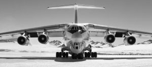 A large cargo airplane is parked on a snowy runway at the South Pole, with Antarctic mountains in the background. The image, captured in black and white, highlights the remote setting of Antarctic Logistics and Expeditions.