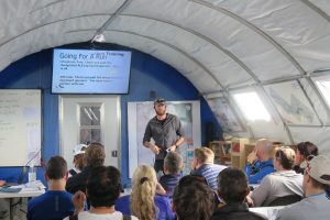A man stands in front of a seated group, giving a presentation inside a large tent in Antarctica. A screen behind him displays Going for a run instructions, and the audience listens attentively in this informal and educational South Pole setting.