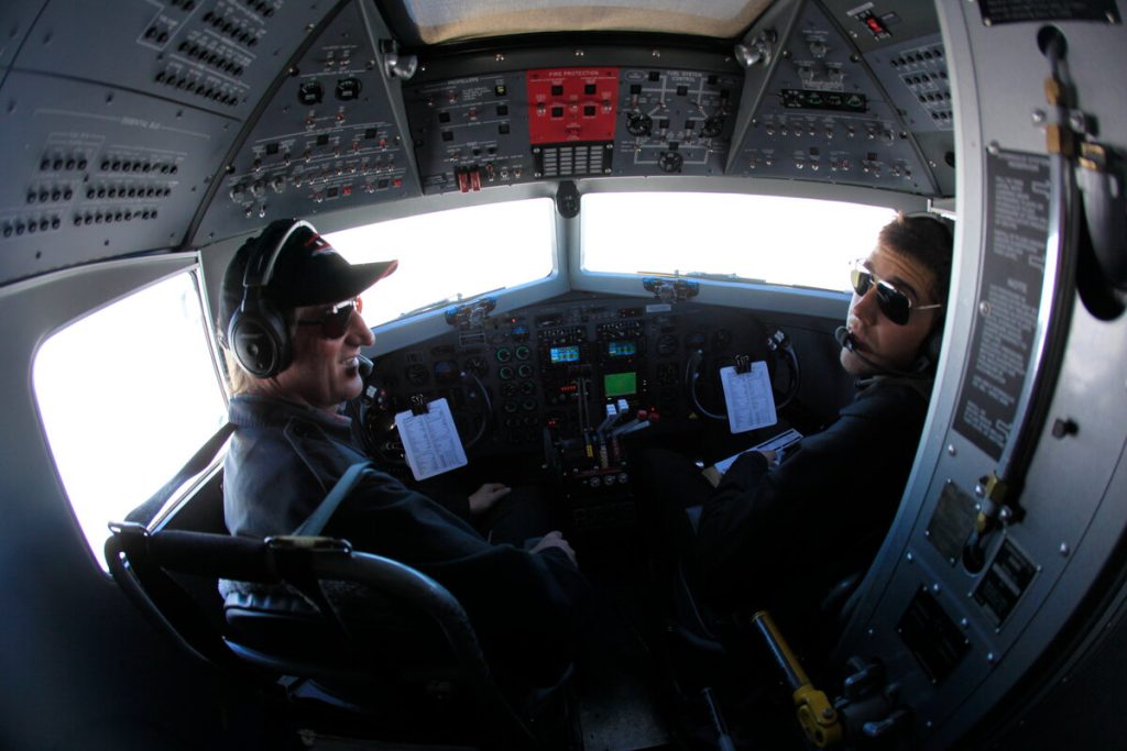 Two pilots wearing headsets and sunglasses sit in the cockpit of an aircraft, surrounded by control panels and instruments, en route to the South Pole with brilliant Antarctic light streaming through the windshield.