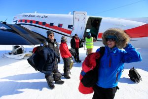 A group of people in winter clothing stand on snow near a small plane with Kenn Borek Air Ltd. written on its side, preparing for or finishing a journey to Antarctica in a cold, sunny environment.