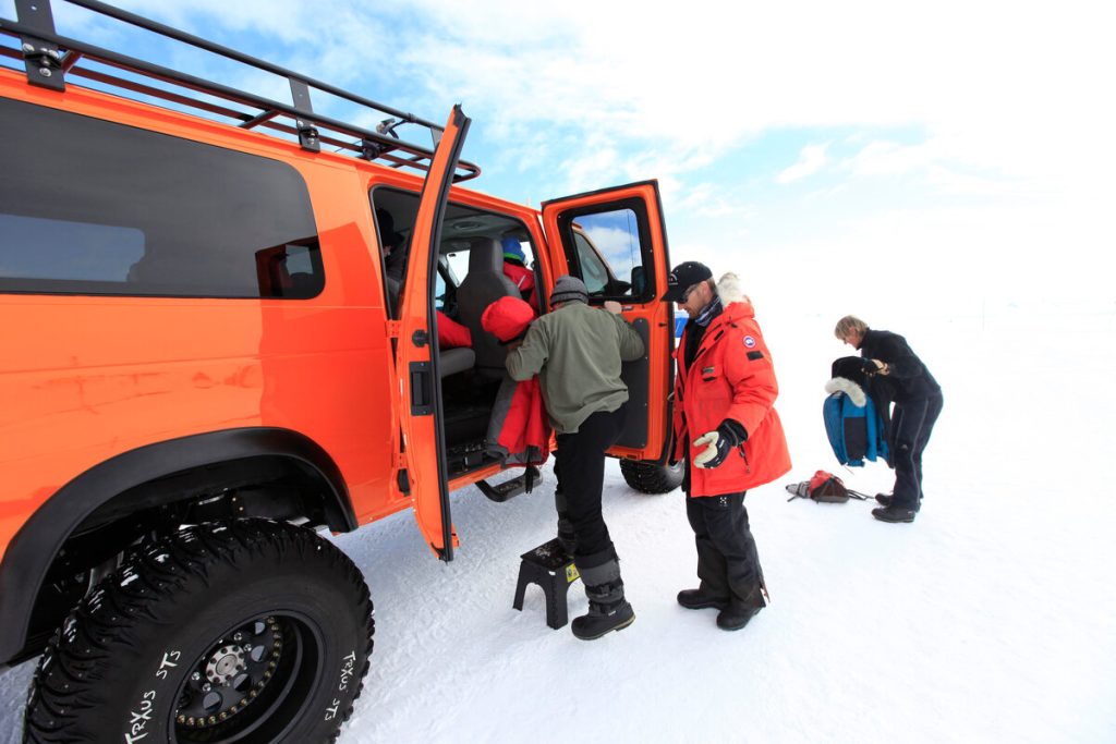 Four people in winter clothing help each other get into a bright orange off-road vehicle on the snow at Antarctica, with one person using a small step stool while another stands nearby holding gear.