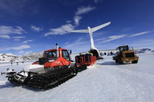 A red snowcat and a yellow loader unload supplies from a large white cargo plane on a snowy, icy runway in Antarctica, with mountains and blue sky in the background—part of Antarctic Logistics and Expeditions at the South Pole.