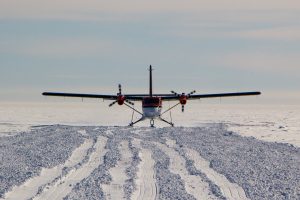 A small twin-propeller airplane faces forward on a snow-covered runway at the South Pole, operated by Antarctic Logistics and Expeditions, with tire tracks in the snow and a vast, icy Antarctic landscape under a cloudy sky.