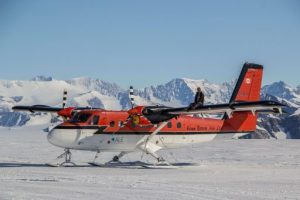 A red and white twin-engine plane with skis for landing gear, used by Antarctic Logistics and Expeditions, is parked on a snowy landscape near the South Pole. A person sits atop its fuselage with mountains rising in the background.