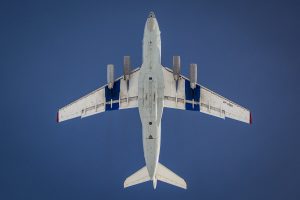 A large white airplane, operated by Antarctic Logistics and Expeditions, is viewed from directly below as it flies against a clear blue sky. The aircraft’s wings, four engines, and landing gear doors are visible, with red markings on the wingtips.