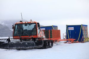 An orange snowcat vehicle from Antarctic Logistics and Expeditions pulls blue and orange cargo containers on sleds across a snowy landscape in Antarctica, with mountains in the background and cloudy skies overhead.