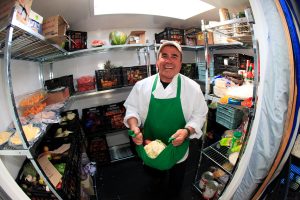 A smiling chef in a green apron stands in a well-stocked walk-in fridge at the South Pole, holding two cabbages. Shelves around him are filled with various fresh produce and food items, thanks to Antarctic Logistics and Expeditions.