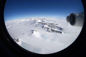 A view through an airplane window shows a snow-covered, mountainous landscape in Antarctica with glaciers and rugged peaks under a clear blue sky. Part of the Antarctic Logistics and Expeditions aircraft’s engine is visible on the right side of the window frame.