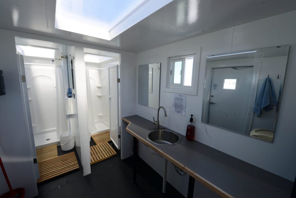 A clean, modern restroom at Antarctic Logistics and Expeditions features a shower area, round-basin sink, long countertop, mirror, small window, fire extinguisher, and towels hanging near the door—ideal comfort even in Antarctica.
