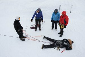 Four people in winter gear stand on snow, handling climbing ropes, while a fifth person lies on the ground as if being rescued, suggesting a South Pole mountaineering or Antarctic Logistics and Expeditions training scenario.