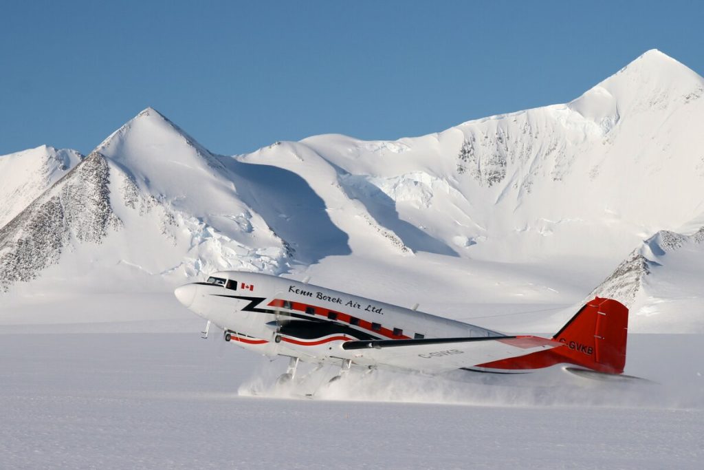 A red and white propeller plane lands on snow in a mountainous Antarctic landscape with clear blue sky, sending up powder near large, jagged, snow-covered peaks—an iconic scene from Antarctic Logistics and Expeditions.