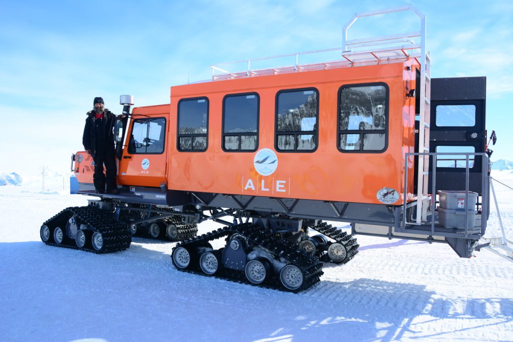 A large orange tracked vehicle with the Antarctic Logistics and Expeditions (ALE) logo is parked on snow, with a person in dark winter clothing standing on its side step. Snowy mountains and the bright sky of Antarctica are in the background.