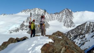 Two climbers with helmets and skis on their backpacks stand on a snowy mountain ridge in Antarctica, surrounded by rugged, snow-covered peaks under a clear blue sky—an adventure made possible by Antarctic Logistics and Expeditions.