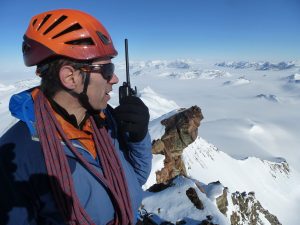 A climber wearing an orange helmet and sunglasses uses a walkie-talkie while standing on a snowy mountain peak in Antarctica, with snow-covered mountains and a vast, clear sky in the background.