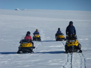 Five people ride yellow snowmobiles across a vast, snowy landscape in Antarctica under a clear blue sky, leaving tracks behind them. All are wearing winter gear and helmets, typical of Antarctic Logistics and Expeditions travelers.