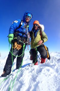 Two climbers in heavy winter gear stand on a snowy mountain summit in Antarctica, smiling at the camera. Surrounded by climbing equipment and ropes, they enjoy clear blue skies and snowy peaks—an adventure with Antarctic Logistics and Expeditions.