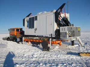 A tracked orange vehicle and two people work with a white portable building on a sled in Antarctica's snowy, icy landscape under a clear blue sky, showcasing the vital work of Antarctic Logistics and Expeditions.