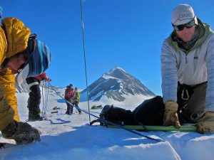Several people in winter gear set up equipment on a snowy mountain slope under a clear blue sky, likely preparing for an Antarctic Logistics and Expeditions journey. A prominent, snow-capped peak rises in the background as one person kneels to adjust gear.