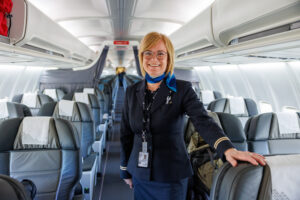 A smiling flight attendant in uniform stands inside an empty airplane cabin, resting one hand on a seat. The bright, clean interior suggests readiness for an Antarctic Logistics and Expeditions flight to the South Pole.